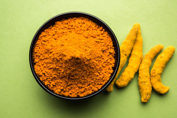 Turmeric powder in ceramic bowl with raw dried turmeric over plain background

