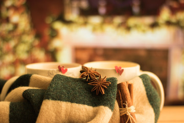 Warm knitted striped scarf is around pair of cups with decoration of red hearts & Christmas spices at against a festive lighting room with beautiful atmosphere of winter holidays. Holidays together.