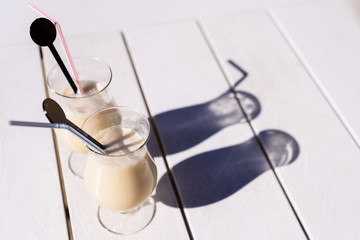 Two glasses of Pina Colada with straws and drinks stirrers on a patio table in sunshine.