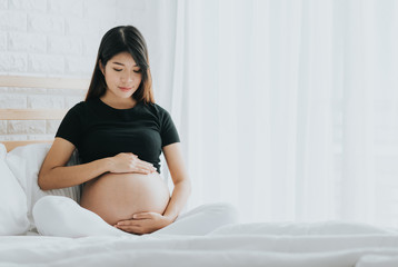 Happy Asian pregnant woman touching her belly with care in bedroom.