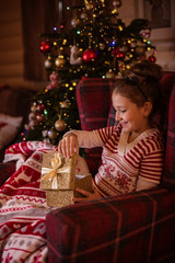 girl sitting in a chair next to a christmas tree opening a present and smiling