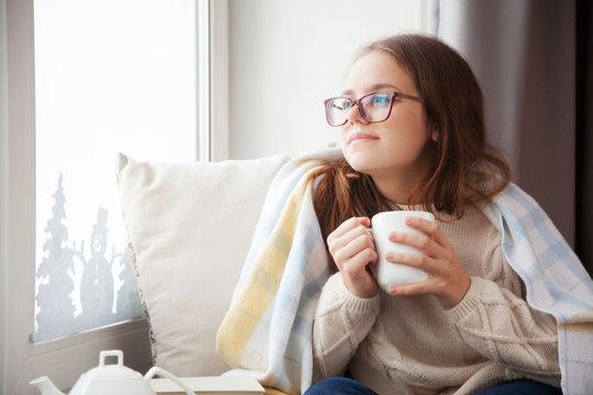 The girl is sitting near the window and holding a white mug. Blankets, blankets and pillows.