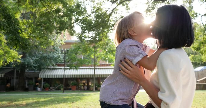 young mother kisses son in lips in backlight at park on weekend