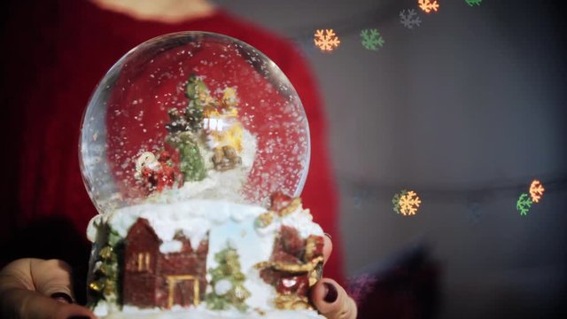 cinemagraph, clouseup hands holding magic globe with snow and santa, on background colorful garland with snowflake figure bokeh, Looping Motion Photo.