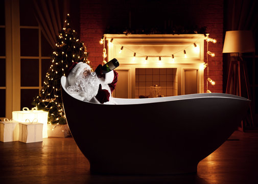 Bad Santa Claus lying in bathtub over christmas tree interior background with retro light bulbs drinking champagne alcohol from the bottle