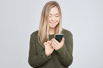 Fototapeta premium .Portrait of pretty young blond Caucasian woman, typing a message to her boyfriend on mobile phone, holding the gadget with one hand, wearing warm wool sweater, looking happy on white wall background.