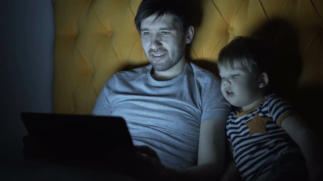 Young father and his little son watching cartoon movie using laptop computer while lying in bed in evening at home