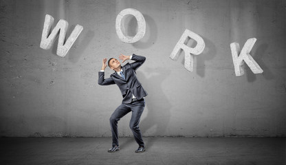 A scared businessman hides under large concrete letters making a word Work above him on a grey background.