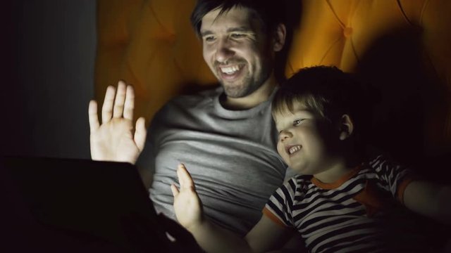 Loving father and his little son having online video chat with grandparents using tablet computer and while lying in bed in evening at home