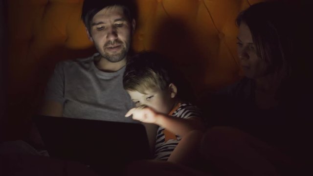 Happy family with little son learning to play tablet computer lying in bed at home in evening