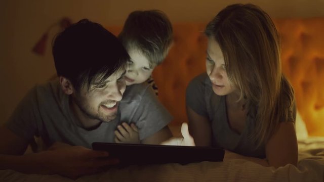 Happy family with little son using tablet computer and talking lying in bed at home before sleeping