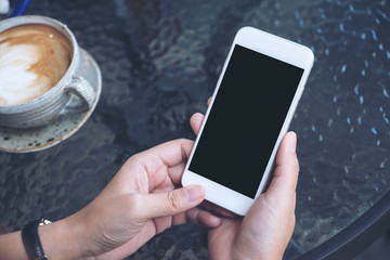 Mockup image of a hand holding white mobile phone with blank black screen and coffee cup on glass table in cafe