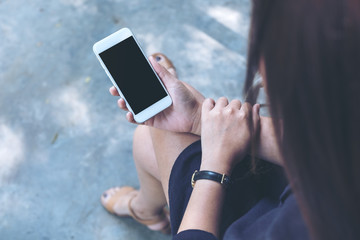 Mockup image of a woman sitting crossed leg and holding white mobile phone with blank black screen with concrete floor background