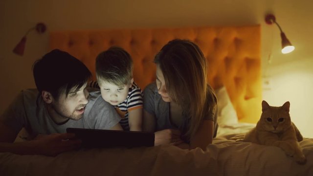 Happy family with little son and funny cat lying in bed at home and using tablet computer for watching cartoon movie before sleeping