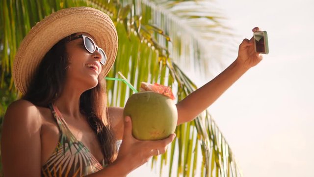 Young Happy Mixed Race Tourist Girl Taking Selfie Using Mobile Phone and Drinking Coconut Cocktail on Tropical Beach. 4K, Slowmotion. Phuket, Thailand.