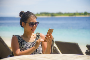 relaxed Asian Chinese woman using internet social media on mobile phone sending text sitting at island vacation resort on the beach