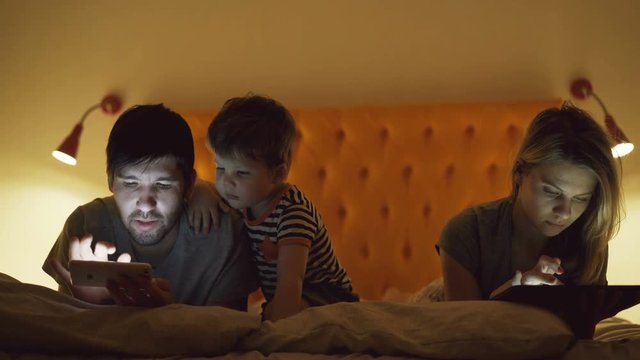 Happy family with little son lying in bed at home using tablet computer and smartphone for watching movie and cartoon in evening before sleeping