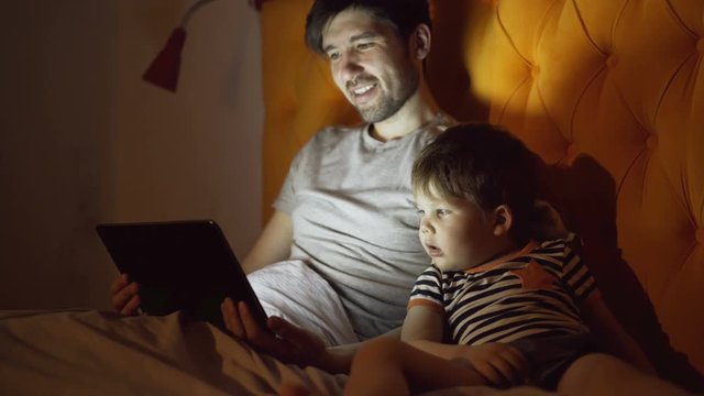 Young father and his little son watching cartoon movie using tablet computer while lying in bed in evening at home