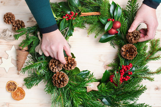 Hands arranging and decorating handmade Christmas wreath with fir branches, pine cones, berries, baubles on the natural wooden table, top vew, selective focus