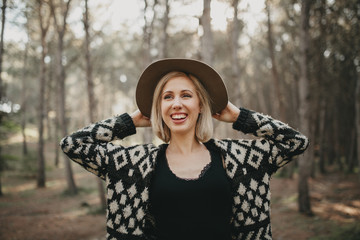 Portrait of blonde white smiling woman putting on a hat in the forest.