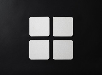 Four square blank white beer coasters on black paper background. Template for your design. Flat lay.