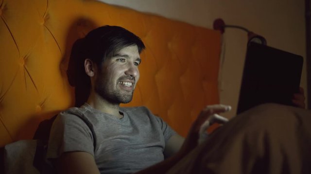 Young laughing man using tablet computer for surfing social media lying in bed at home before sleeping