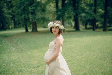 Fototapeta na wymiar Happy pregnant woman in a soft beige dress and a flower wreath on the head walks around the spring park