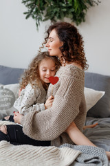 Vertical shot of affectionate young mother and her daugher embrace each other and express love, sit on comfortable bed, close eyes with enjoyment, being relaxed and pleased. Parenthood concept.