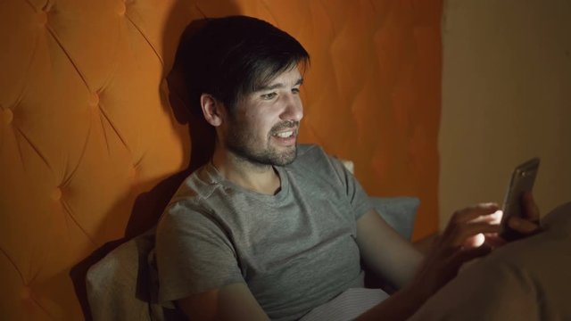 Young smiling man using smartphone for surfing social media lying in bed at home at night