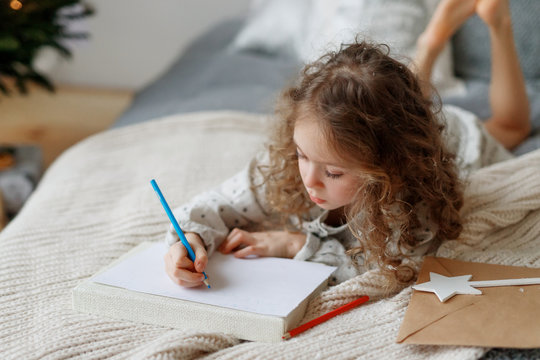 Portait of small lovely beautiful curly child draws pictures on blank white sheet of paper, wants to congratulate her best friend with coming New Year, lies on comfortable bed. Talented female kid.