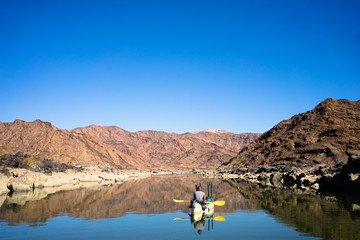 A canoe drifting along the calm waters of the Orange river with rugged mountains reflecting off the calm water - 184801881