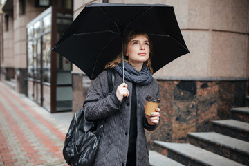 Portrait of young cute lady standing on street with black umbrella and coffee in hands