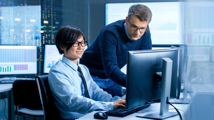 In the Data Mining Center Chief Statistician Consults His Young Assistant sits at His Workstation Surrounded with Monitors Displaying Data and Graphs.