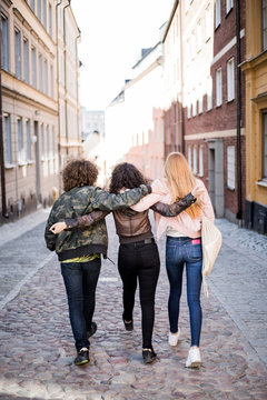 Rear view of friends walking with arms around on cobbled street in city