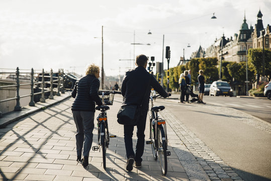 Full length rear view of senior couple with bicycles walking on sidewalk in city