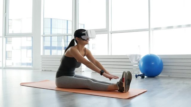 Body training. Full length of attractive young woman doing stretching exercises while sitting on the floor and looking through virtual glasses