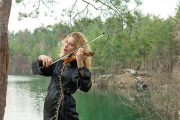 A beautiful young girl plays the violin on the shore of the lake.