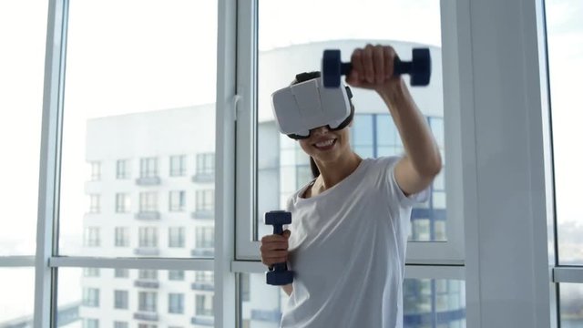Entertainment. Close up of cheerful woman doing boxing movements while holding light weights and wearing virtual reality glasses