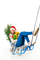 Obraz premium laughing girl in santa hat sitting on sled with gifts and christmas tree, isolated on white