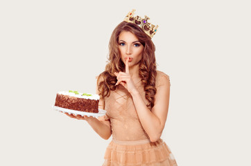 Shh it's a surprise. Woman holding a cake in her hand and showing silence quiet sign gesture isolated white background. Girl full makeup curly brown long  hair blue eyes lace dress party crown on head