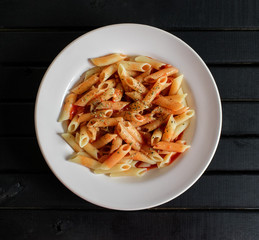 Cooked Penne Pasta Dish, with vibrant red tomato sauce on top, and sprinkled oregano, on a white porcelain plate, black dark wood background tabletop