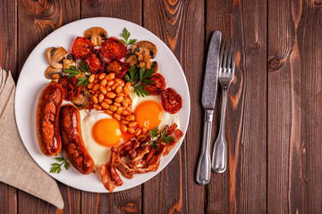 Traditional full English breakfast with fried eggs, sausages, beans, mushrooms, grilled tomatoes and bacon on wooden background.