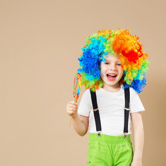 Happy clown boy in large colorful wig. Let's party! Funny kid clown. 1 April Fool's day concept. Portrait of a child eating lollipop.