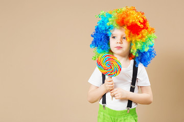 Happy clown boy in large colorful wig. Let's party! Funny kid clown. 1 April Fool's day concept. Portrait of a child eating lollipop.