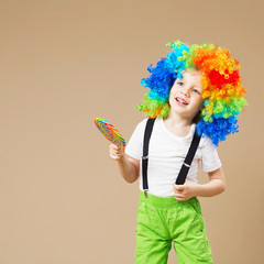 Happy clown boy in large colorful wig. Let's party! Funny kid clown. 1 April Fool's day concept. Portrait of a child eating lollipop.
