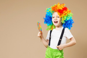 Happy clown boy in large colorful wig. Let's party! Funny kid clown. 1 April Fool's day concept. Portrait of a child eating lollipop.