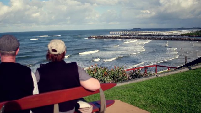 a couple sitting on bench looking at surfers catching wave on ocean
