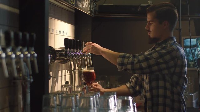 The side view of the barman with dark beard and the brooklyn bartender mustaches pouring the light beer into the glass using the draft beer tower at the background of the bar.