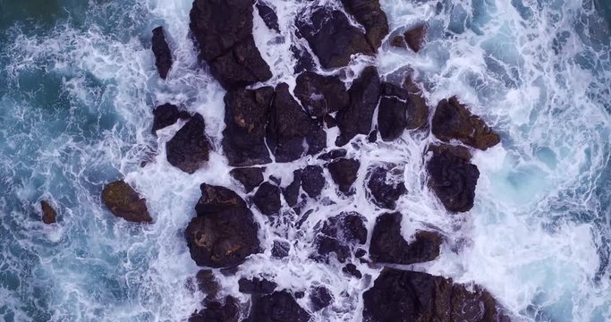 Top down aerial shot of white water and turquoise waves washing over black lava rocks. 