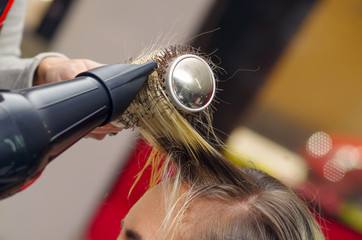 Close up of a hair stylist drying blond hair with hair dryer and round brush in a blurred background